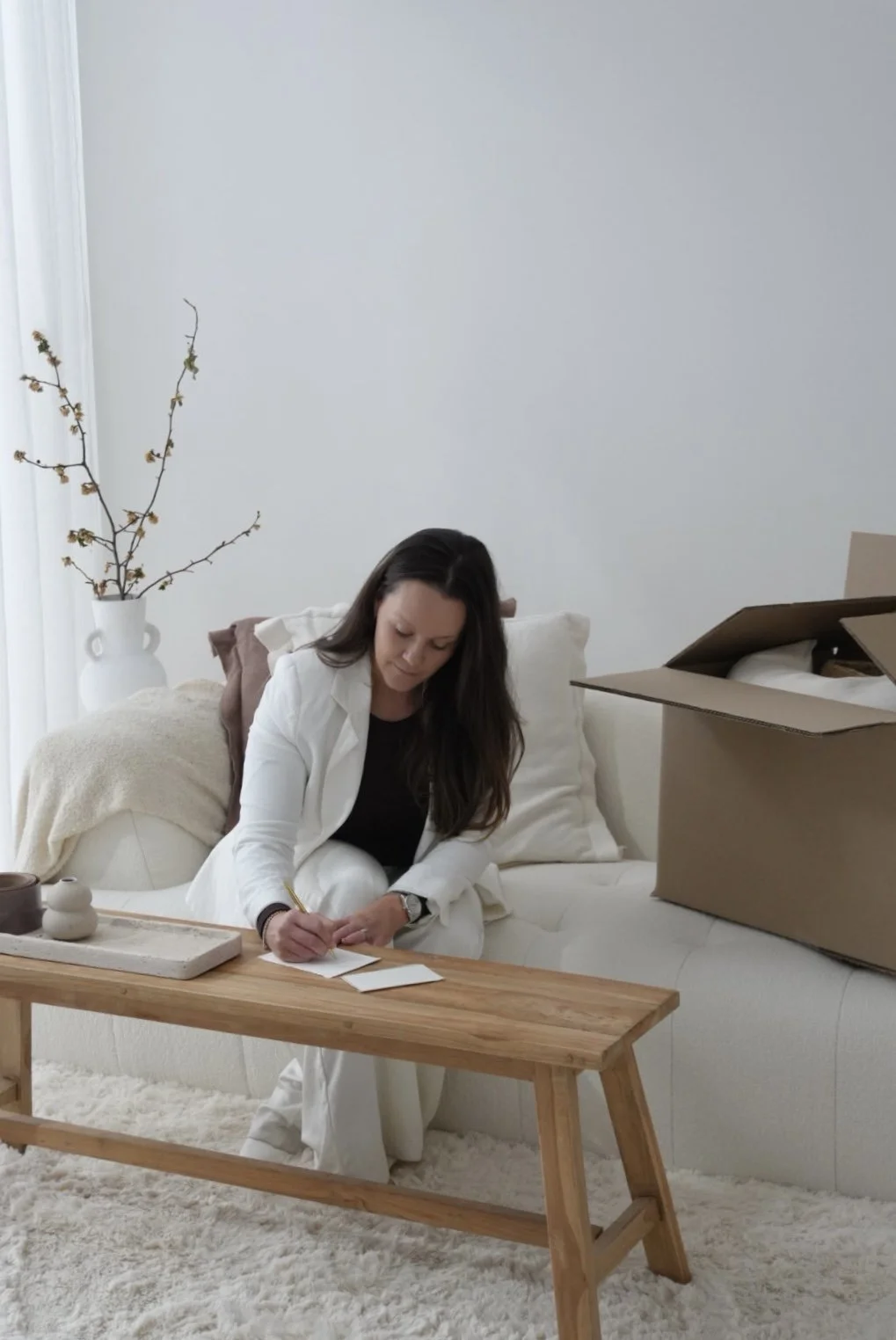 Woman in white clothing writing a note at a wooden coffee table beside an open moving box in a bright living room