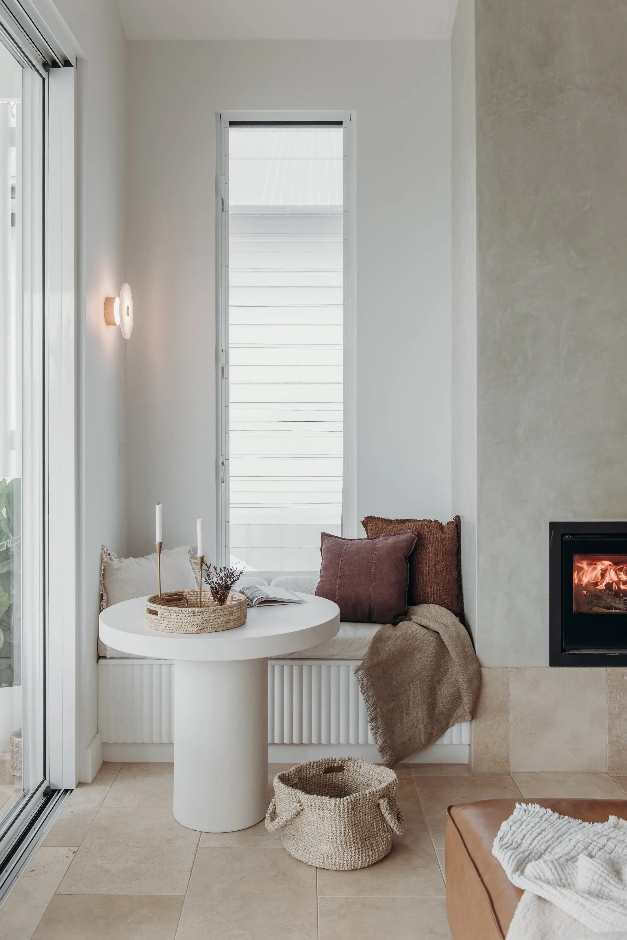 Cozy modern living room with a window seat, white round table, woven baskets, fireplace, cushions, and neutral decor
