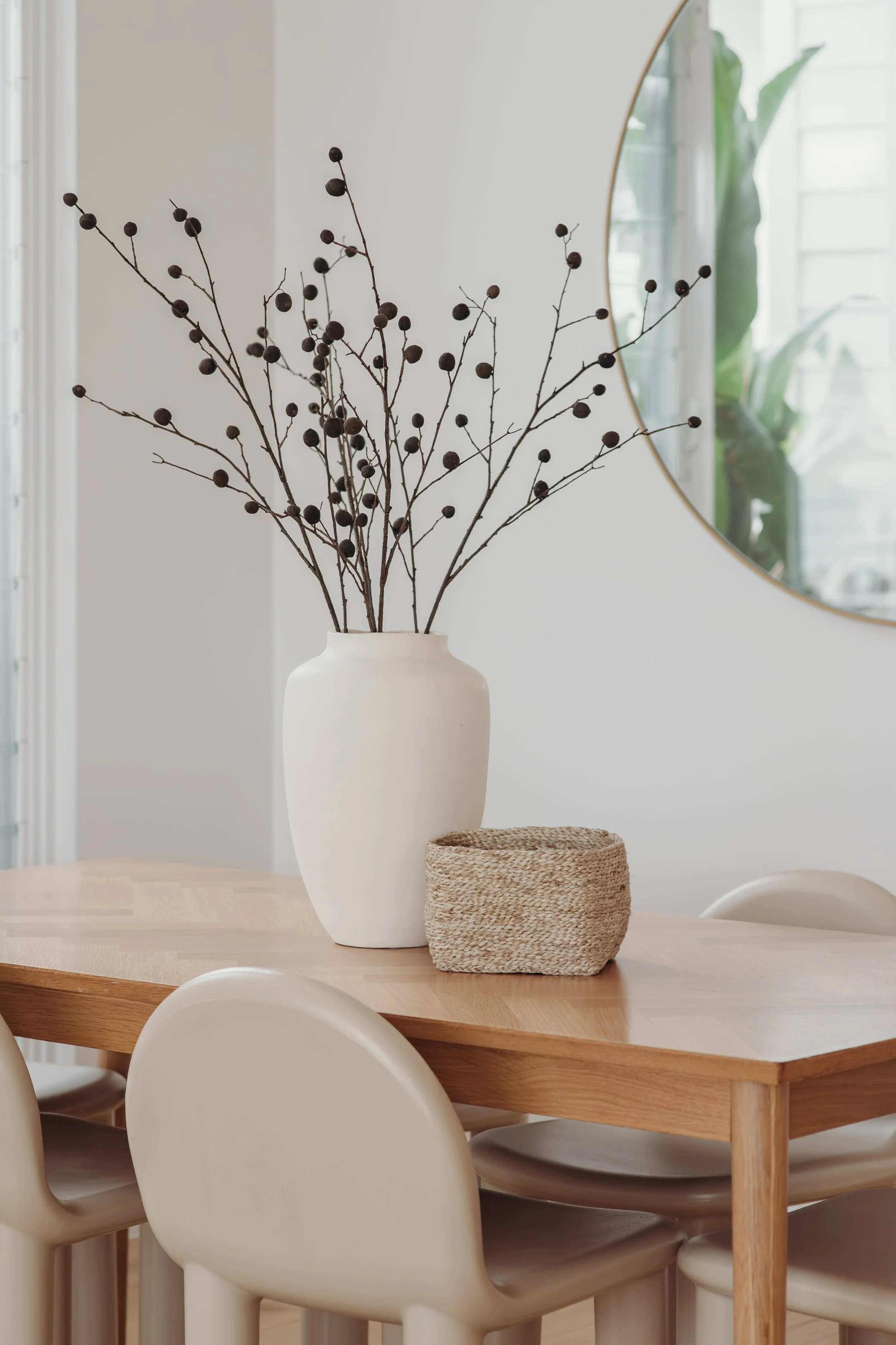 Dining table with a white vase of dried berry branches, woven basket, and modern chairs in a bright room