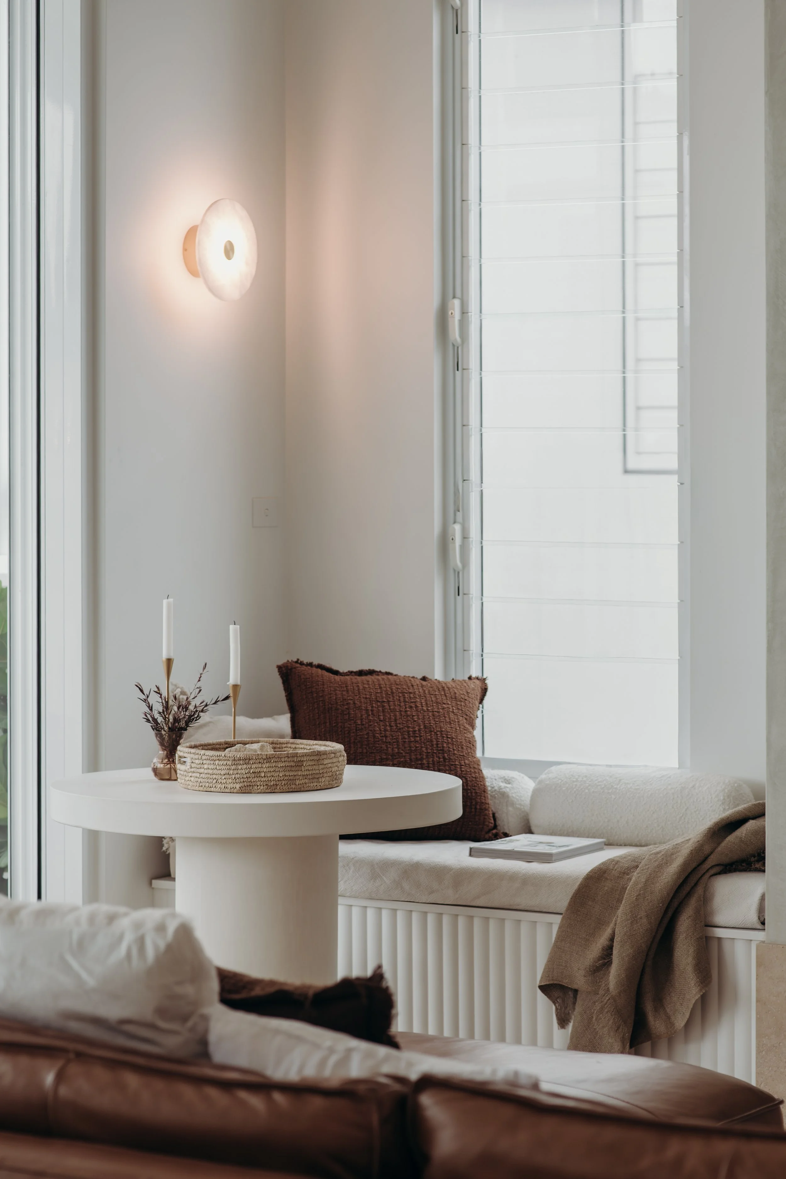 Cozy reading nook with a white built-in window seat, brown pillows, woven basket, candles, and round table
