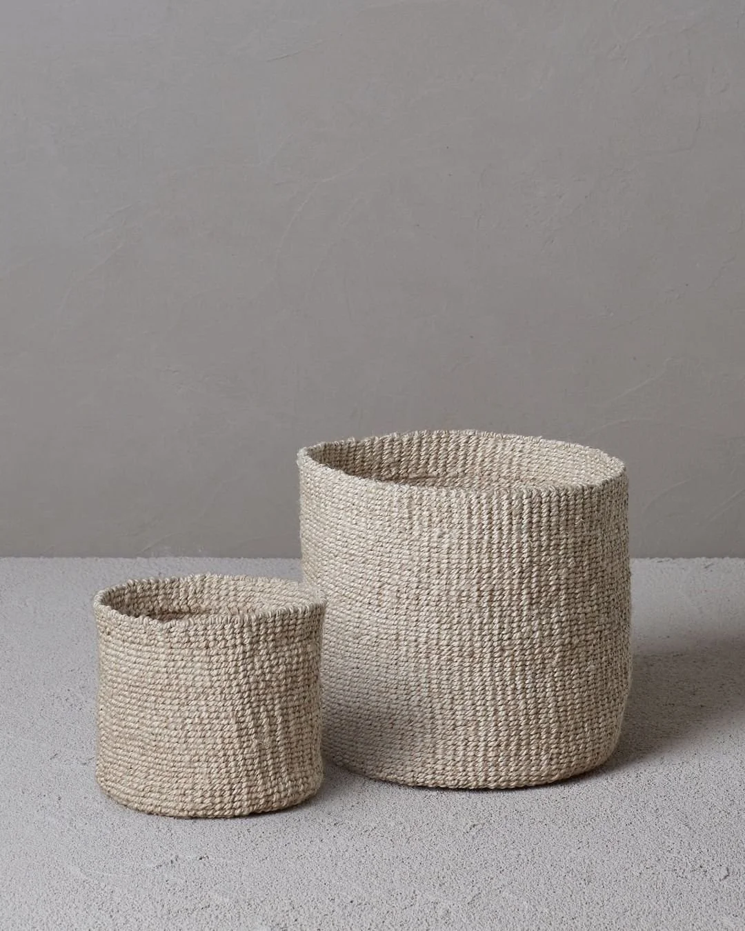 Two natural woven baskets in different sizes on a gray floor against a textured wall