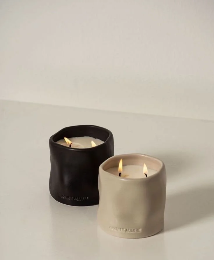 Two lit candles in black and beige ceramic containers on a white surface