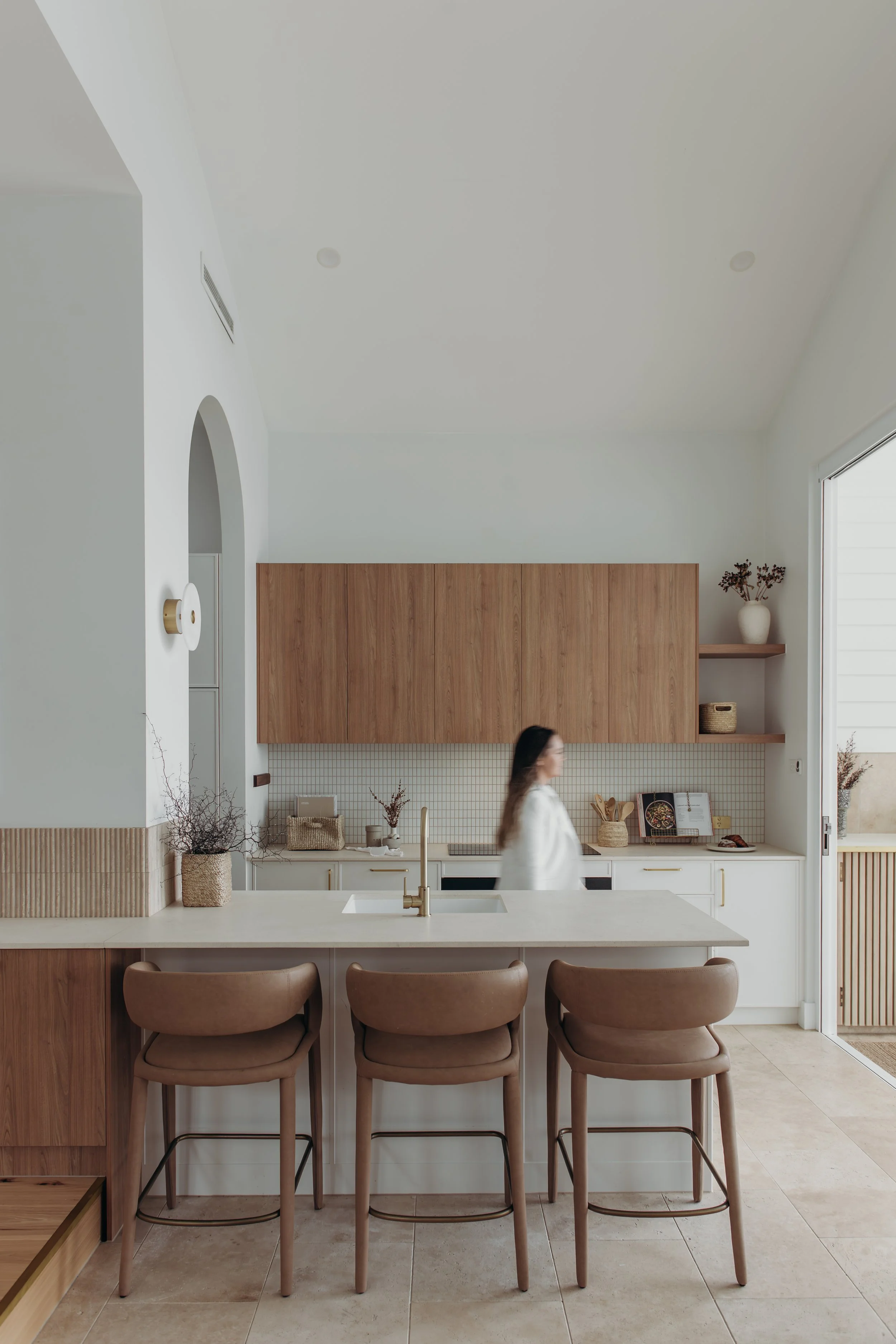 Modern minimalist kitchen with a large island, three leather bar stools, wood cabinetry, tiled backsplash, and a blurred woman walking.