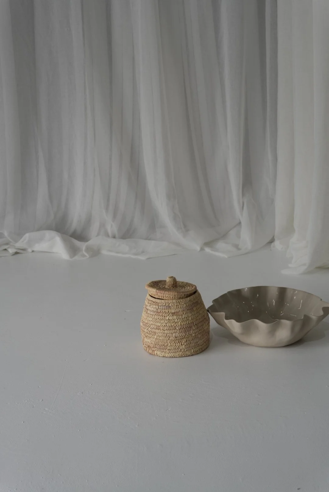Woven lidded basket and wavy ceramic bowl on a minimalist gray floor in front of white curtains