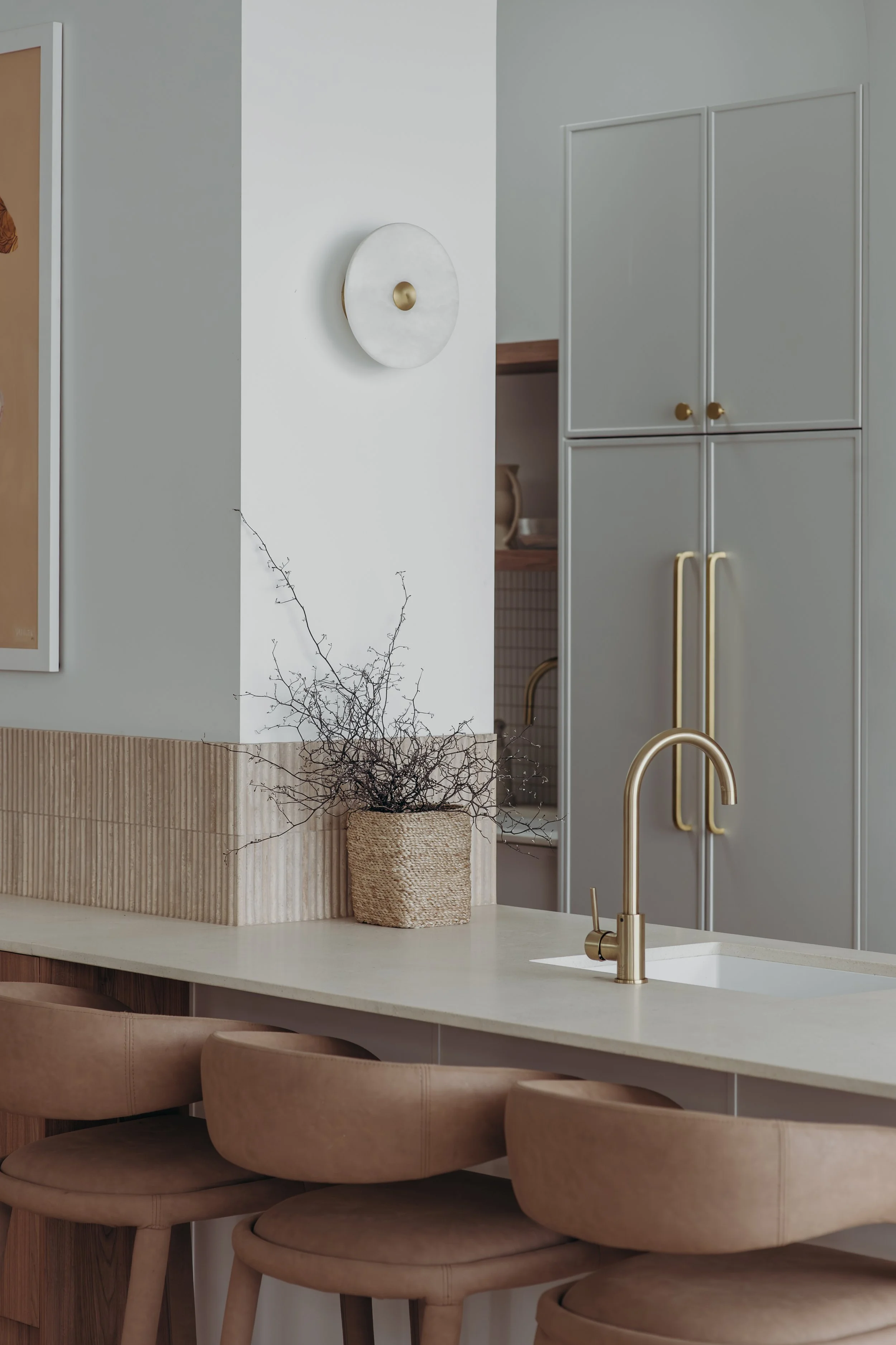 Modern kitchen with white island countertop, gold faucet, gray cabinets, woven basket of branches, and tan bar stools
