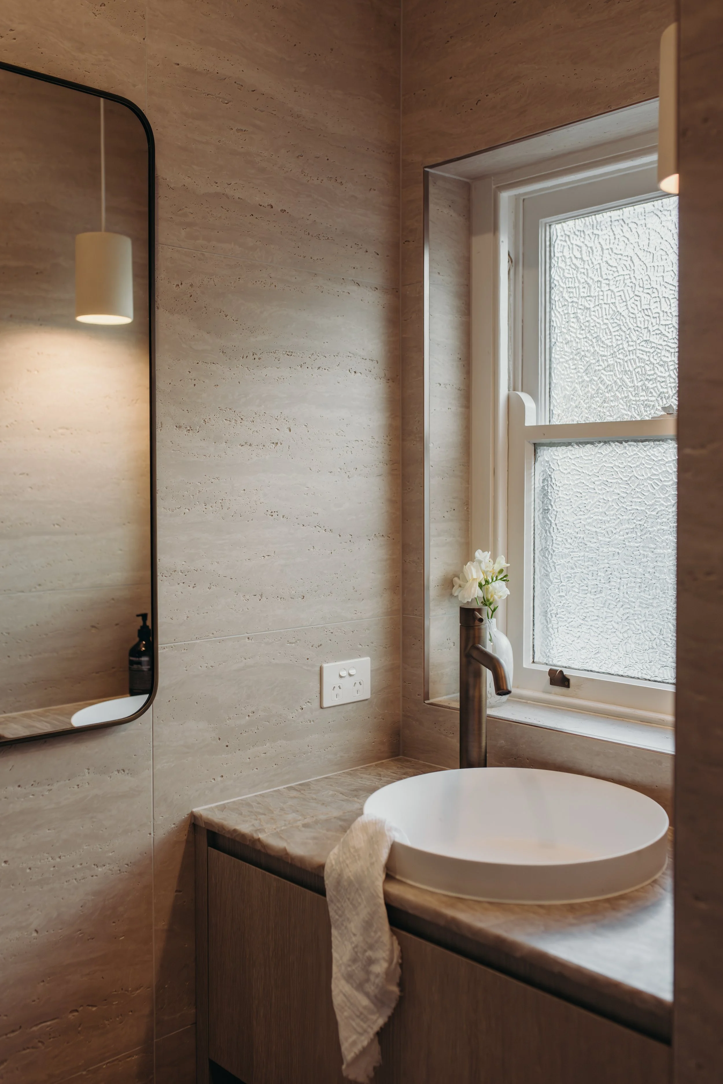 Modern bathroom vanity with white vessel sink, bronze faucet, stone walls, mirror, and frosted glass window