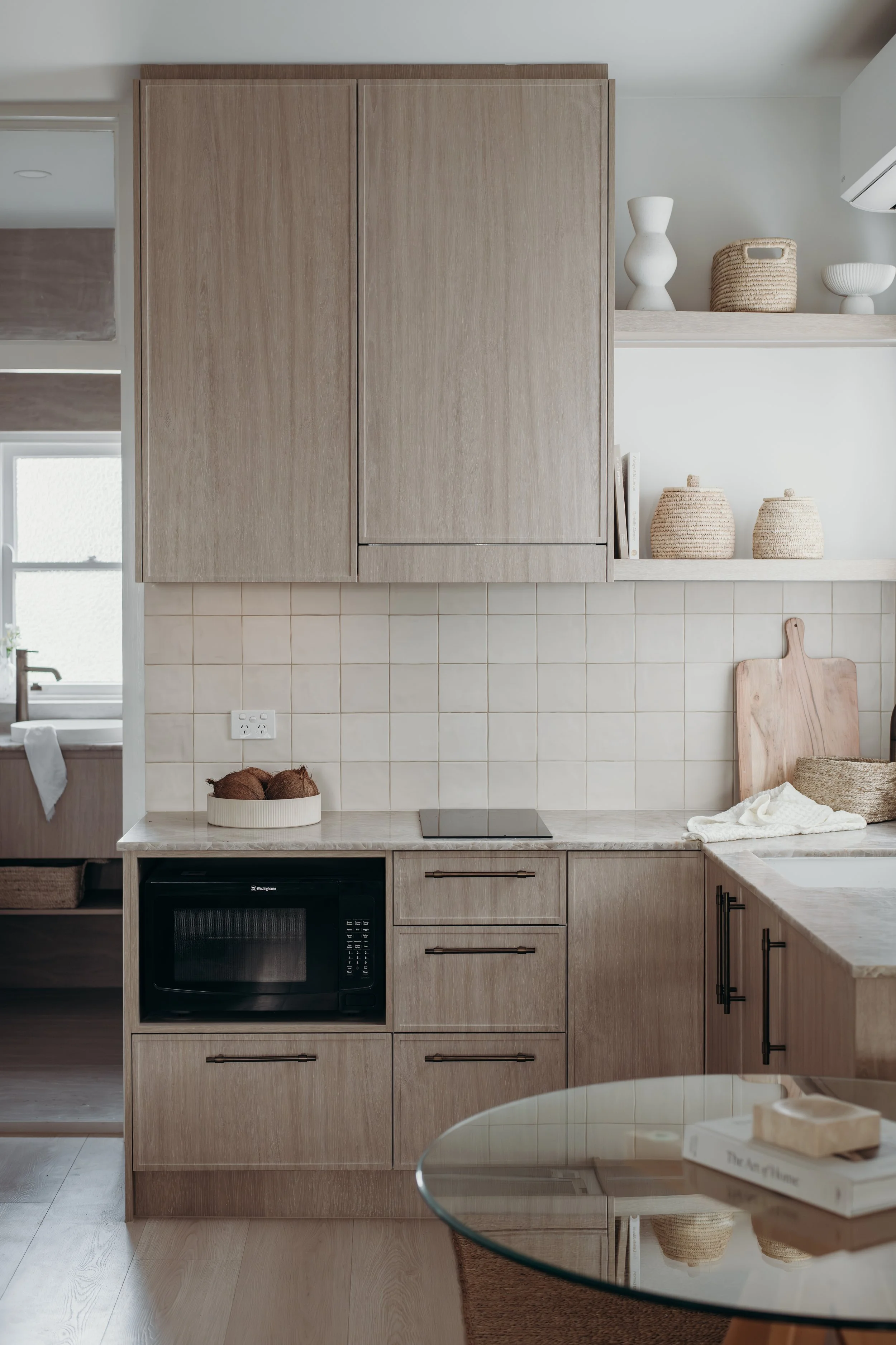 Modern light wood kitchen with tiled backsplash, microwave, cooktop, open shelving, woven baskets, and marble island countertop
