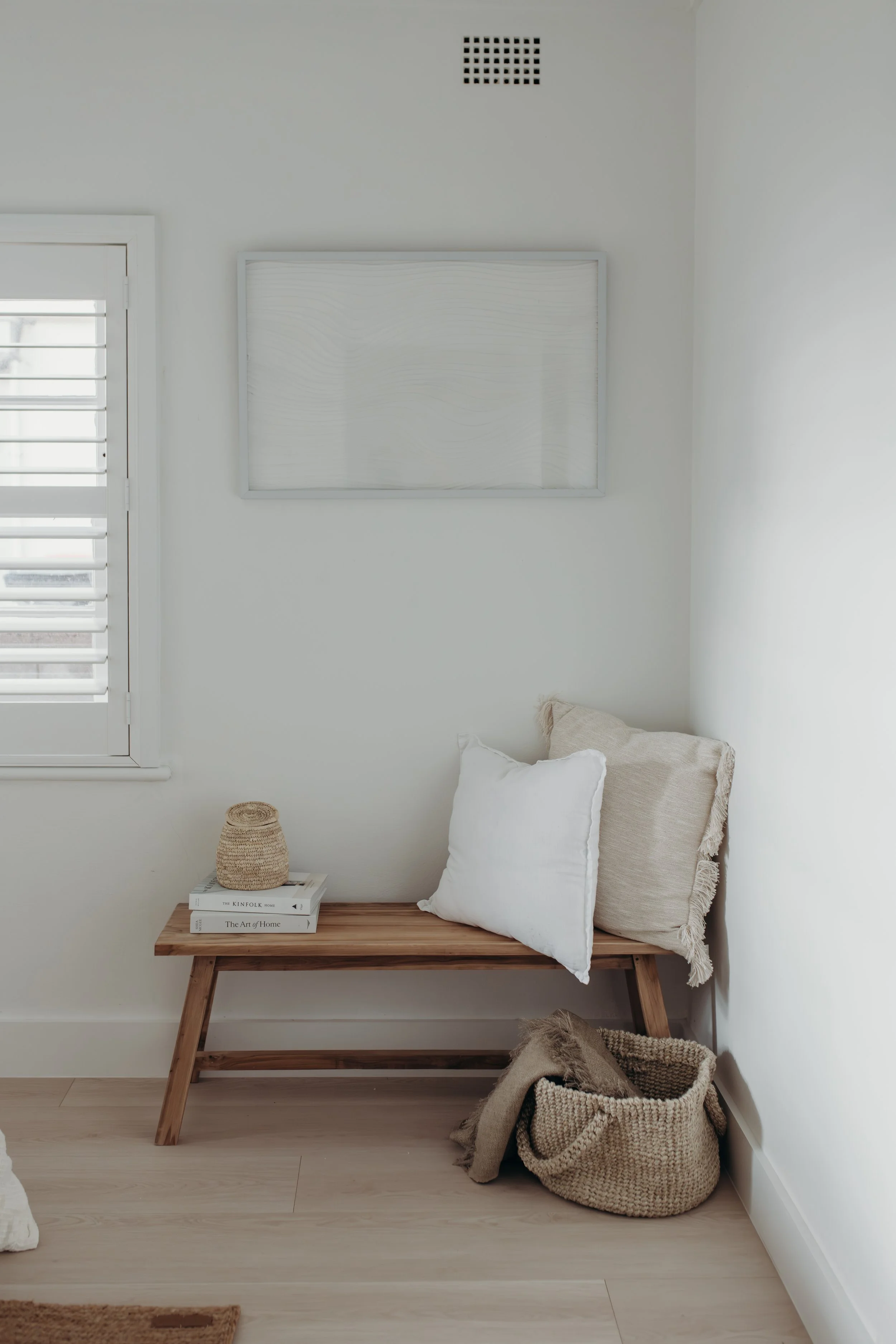 Minimalist bedroom nook with a wooden bench, neutral pillows, woven basket, books, window, and framed wall art