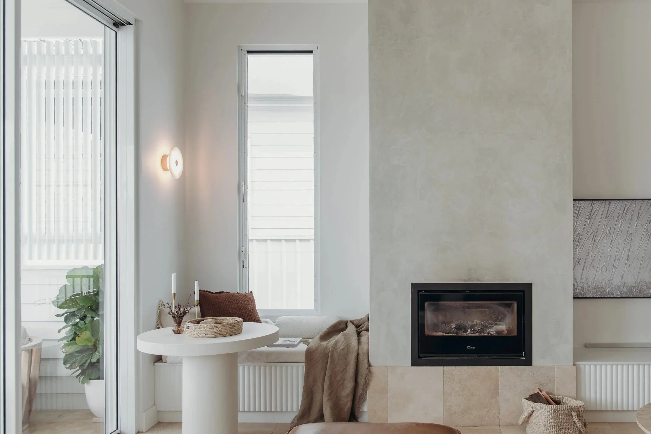 Bright modern living room with a fireplace, round white table, sofa, large windows, and indoor plants