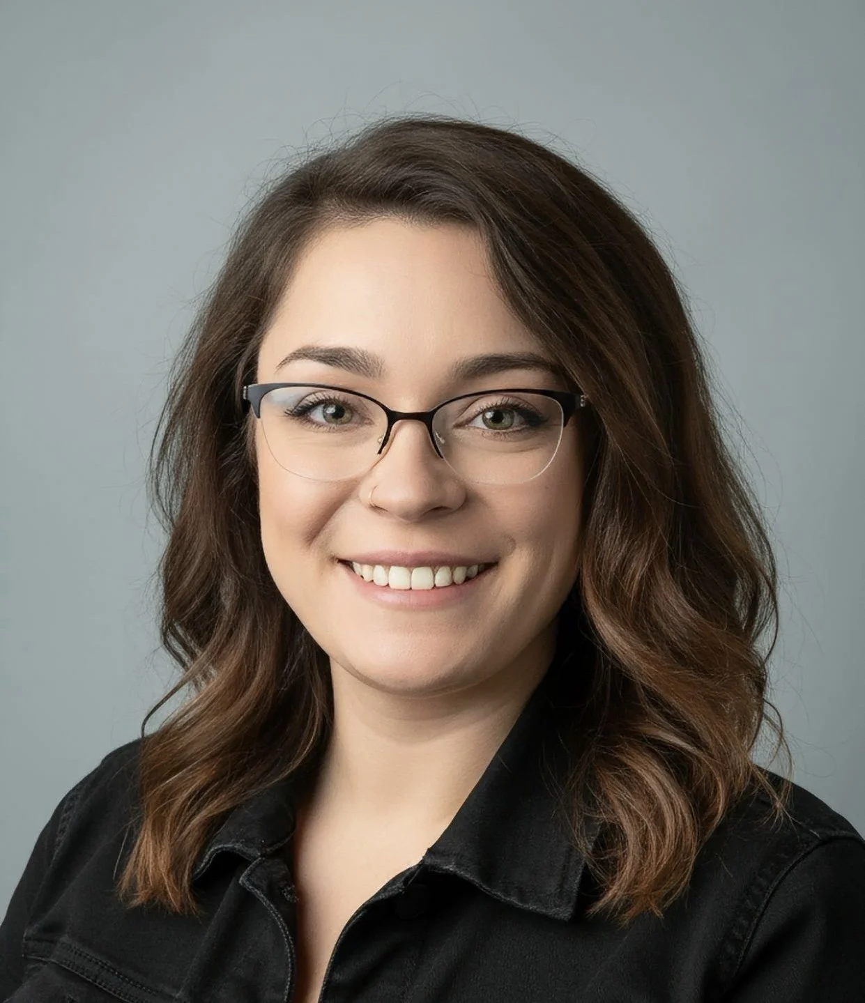 A young woman with shoulder-length wavy brown hair, wearing glasses and a black shirt, smiling against a light gray background.