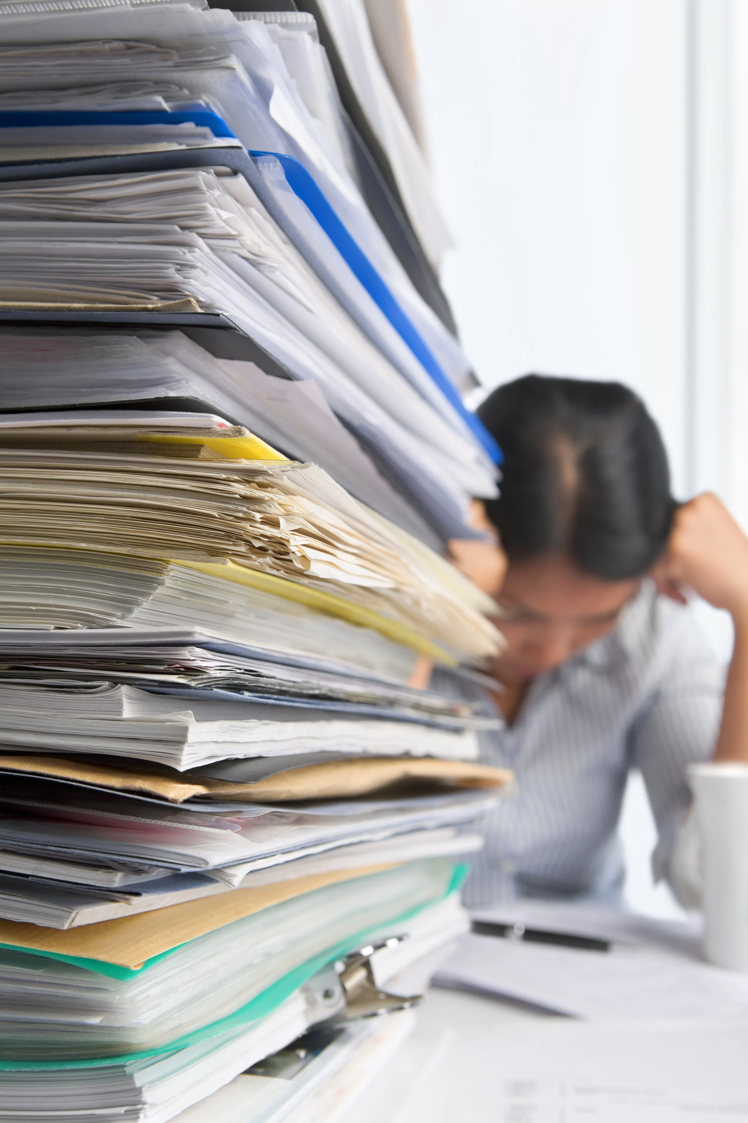 Large stack of disorganized papers and files on a desk with a person blurred in the background.