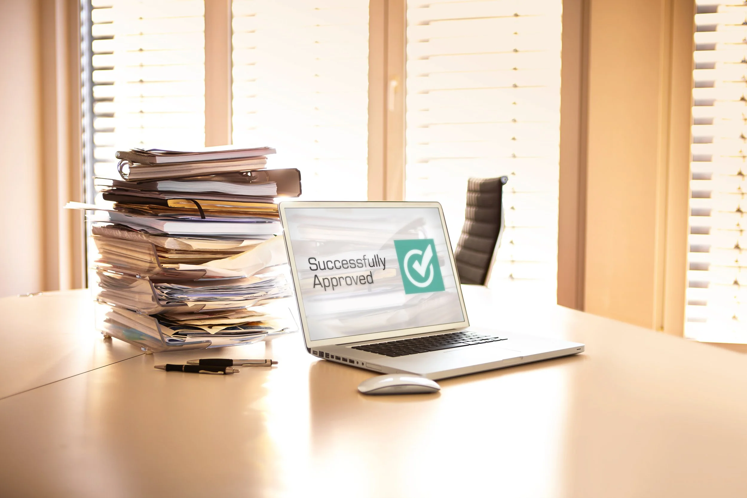 A cluttered office desk with a large stack of documents, a laptop displaying a 'Successfully Approved' message, two pens, and a window with horizontal blinds in the background.
