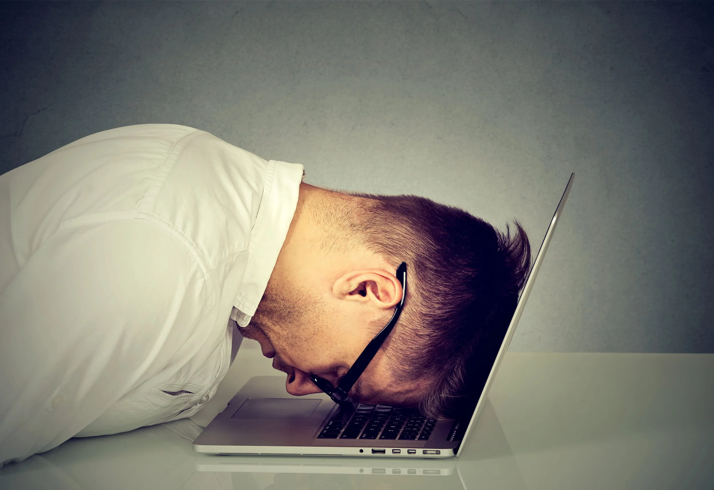Side view of a man wearing glasses, white shirt, working on a laptop in a minimalist space.