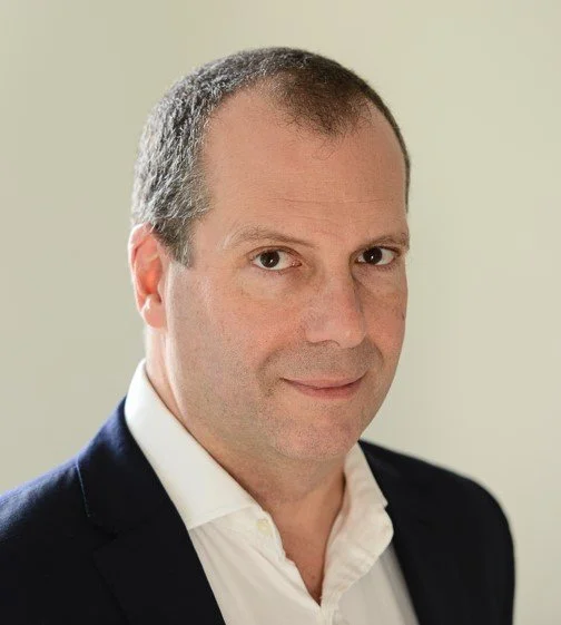 A professional man with short, dark hair, wearing a navy blazer and white shirt, looking at the camera with a neutral expression.