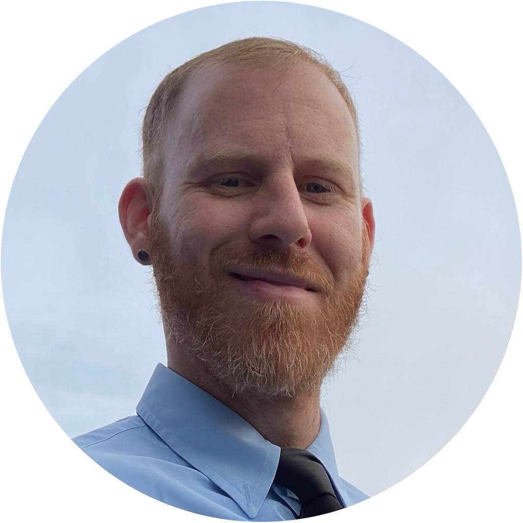 A man with a red beard and short red hair, wearing a blue dress shirt and a black tie, smiling against a light blue sky background.
