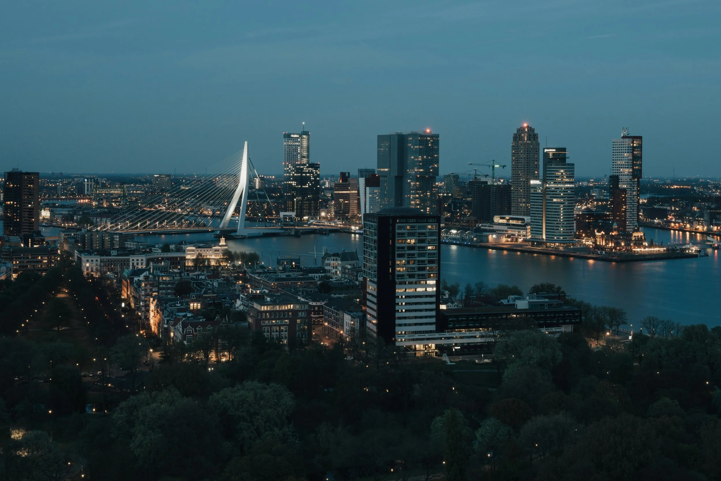 Nighttime aerial view of a city skyline with illuminated skyscrapers, bridges, and a river, under a cloudy sky.