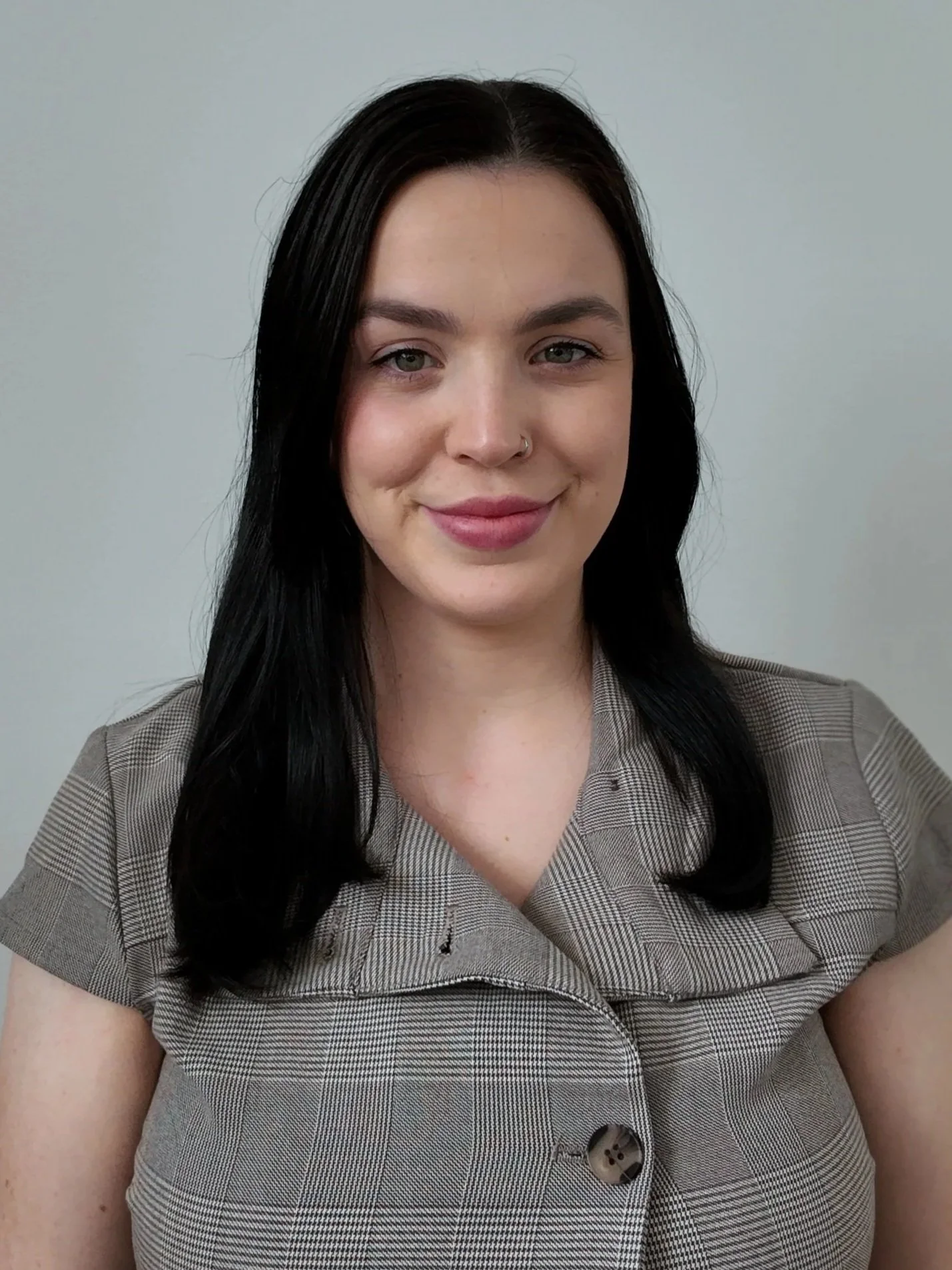 A woman with black hair, fair skin, light makeup, wearing a beige plaid business shirt with short sleeves and buttons.