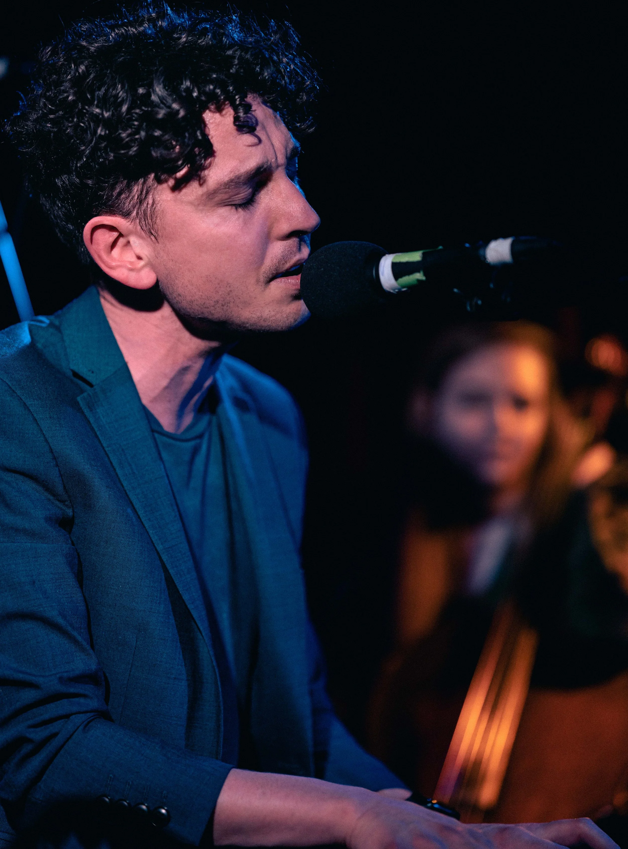 Henry Facey singing into a microphone under dim blue lighting, with his cellist in the background.