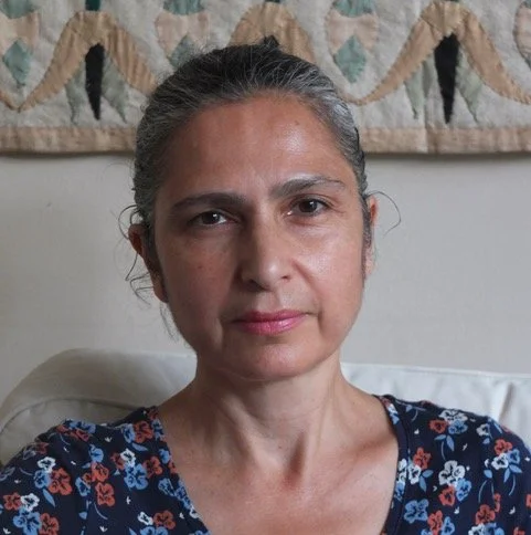 A woman with gray hair pulled back, wearing a dark floral shirt, sitting on a sofa with a decorative quilt hanging on the wall behind her.