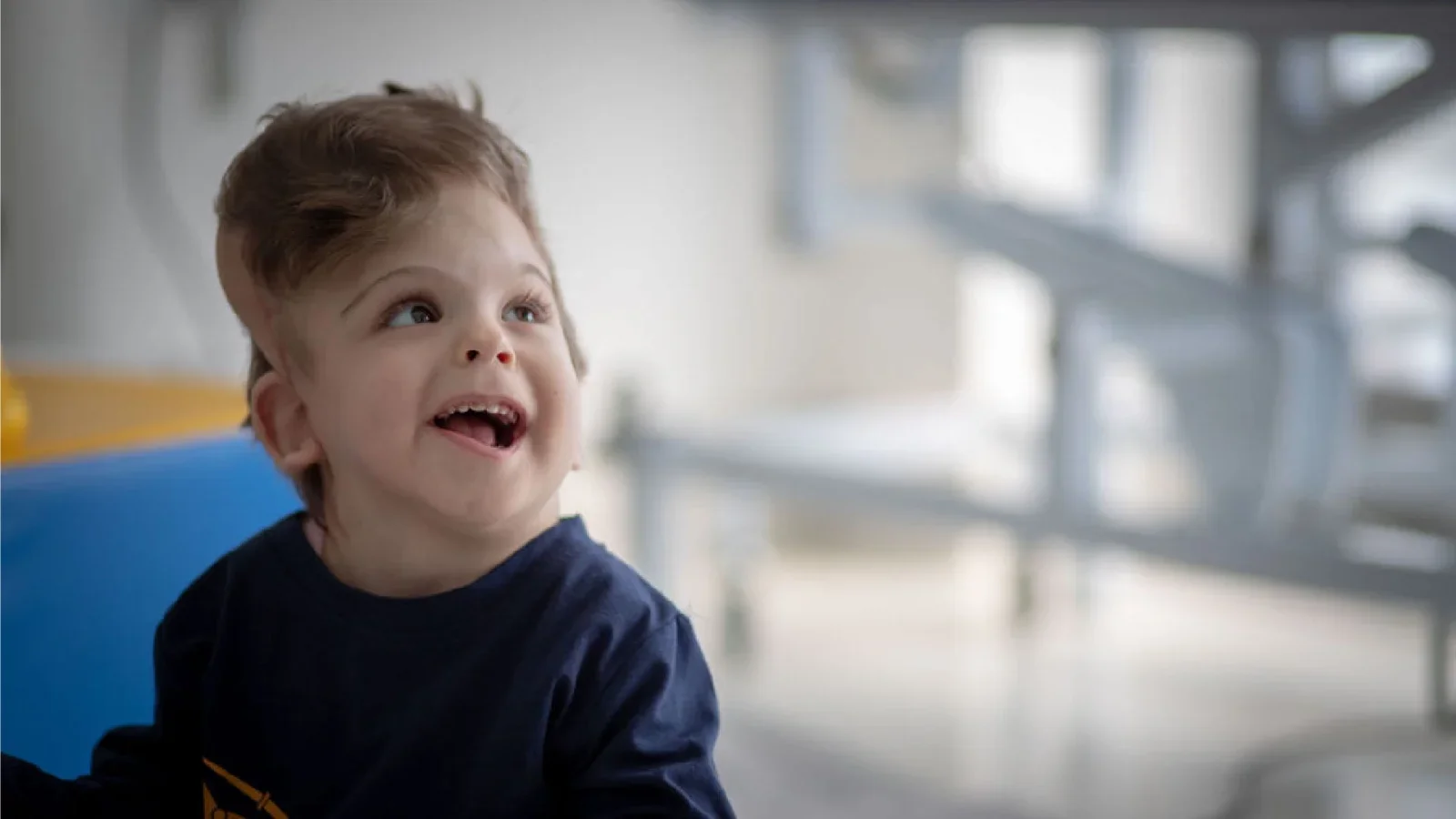 A young boy with brown hair and a blue shirt, smiling and looking upward, seated in a brightly lit room with blurred background.