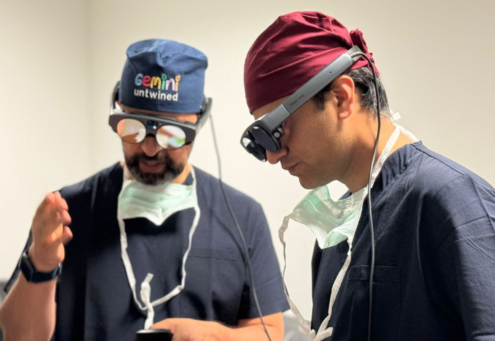 Two medical professionals in scrubs and surgical caps wearing virtual reality headsets, engaged in a training or simulation, with face masks hanging around their necks.