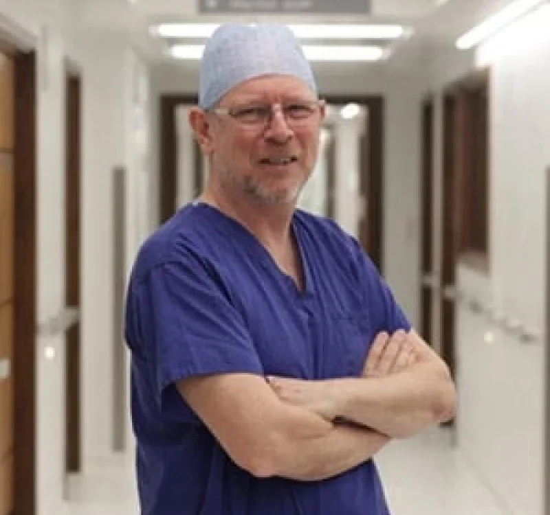 A male healthcare professional in scrubs and a surgical cap standing in a hospital corridor with his arms crossed.