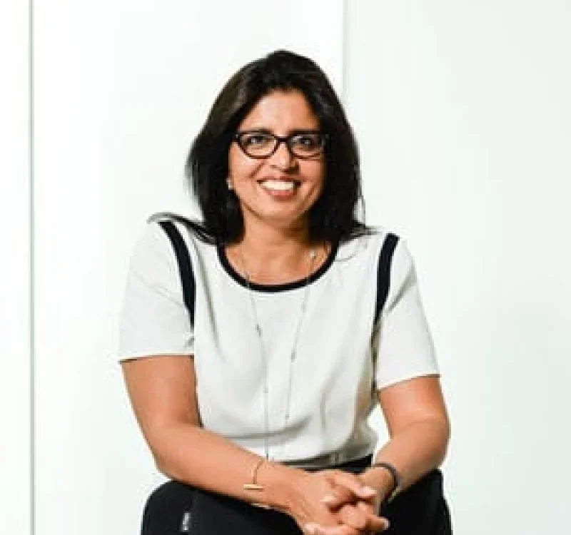 A woman with dark hair, glasses, and wearing a white blouse with black accents, smiling and sitting with her hands clasped against a plain white background.