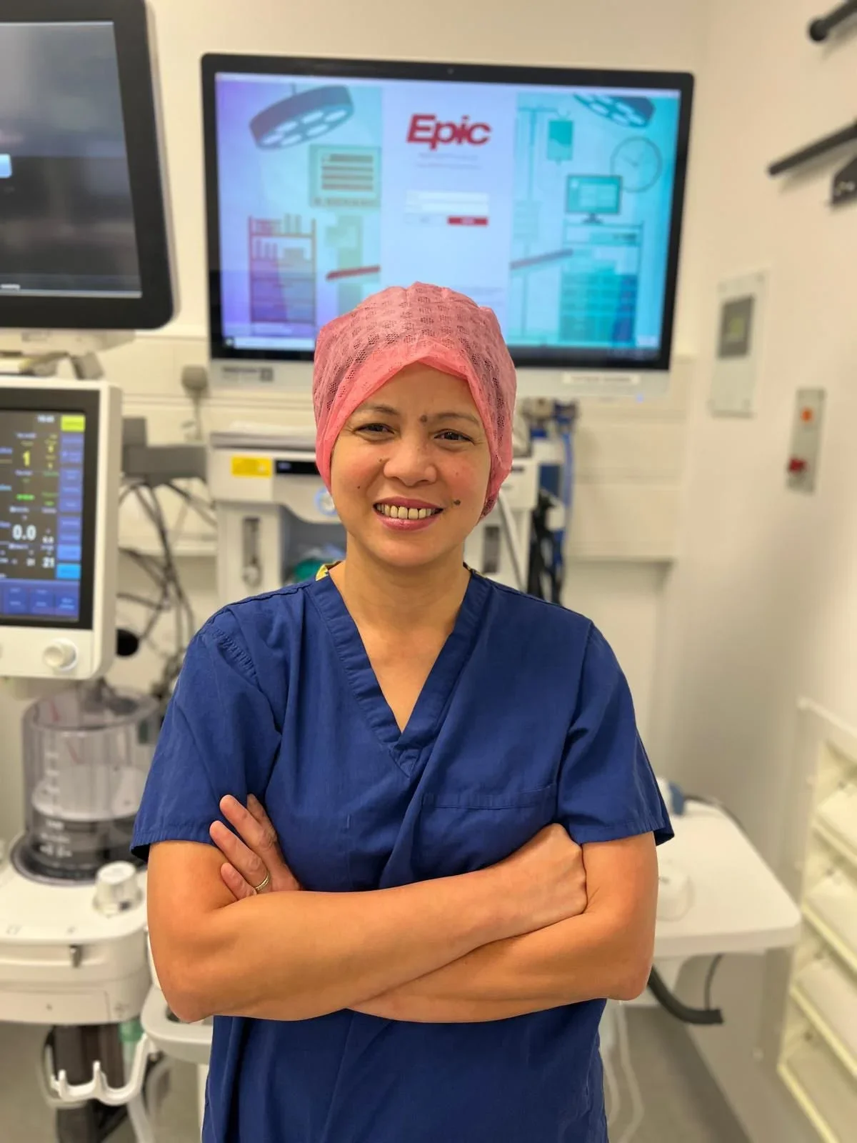 A smiling nurse or medical professional wearing blue scrubs and a pink hair cover standing with arms crossed in a hospital or medical setting with medical equipment and monitors in the background.