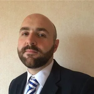 A man with a beard and bald head wearing a dark suit and a white shirt with a blue striped tie, standing against a plain beige wall.