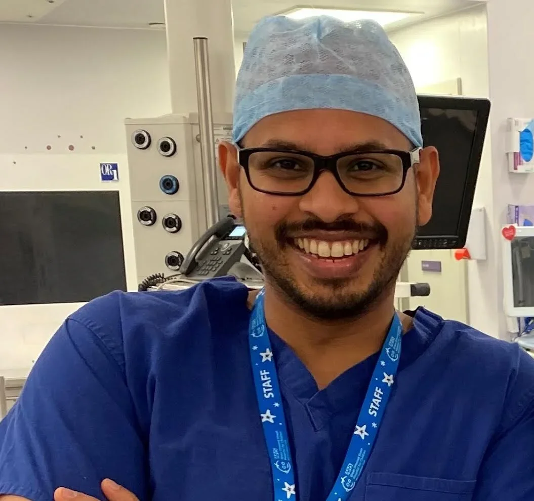 A smiling male healthcare professional wearing blue scrubs, a blue surgical cap, and glasses in a hospital or medical setting.