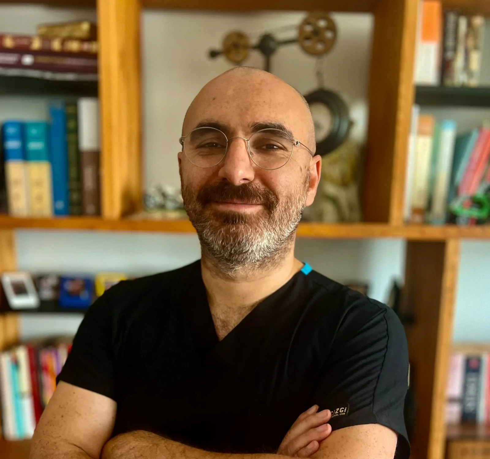 A bald man with glasses and a beard smiling, wearing a black shirt, standing in front of a wooden bookshelf filled with books and decorative items.