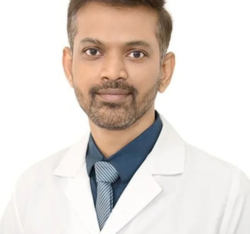 Portrait of a man in a white medical coat, wearing a dark shirt and tie, with short dark hair and a beard, against a white background.
