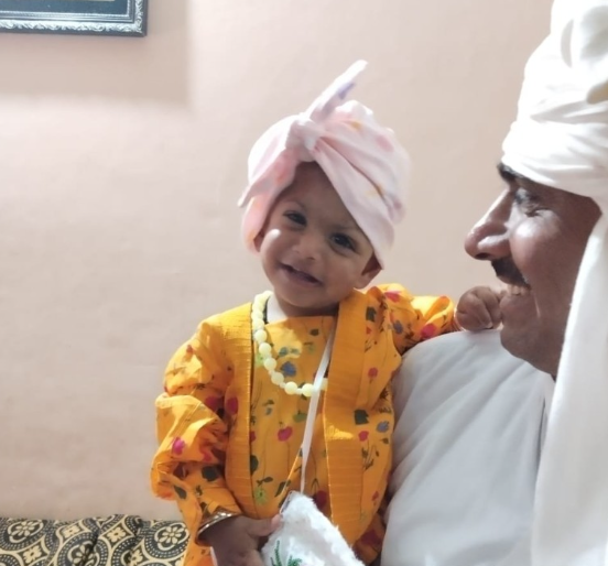 A young girl with a pink headwrap, wearing a bright yellow and orange patterned outfit, smiling while leaning on an older man dressed in white with a white turban, in an indoor setting.