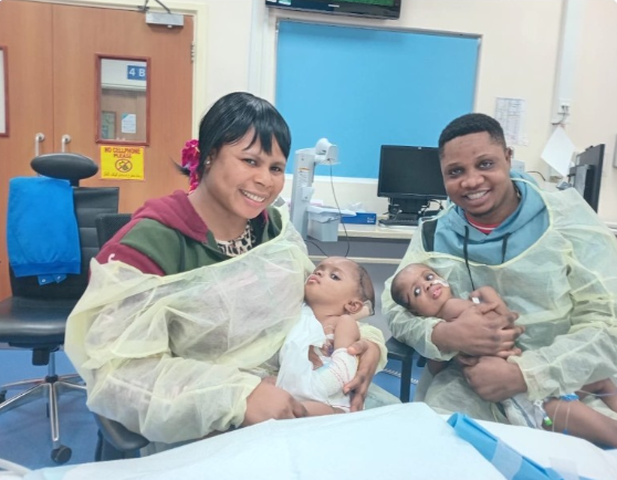 A woman and man at a hospital with two babies, all wearing protective gowns, in a medical room with equipment and a blue wall.