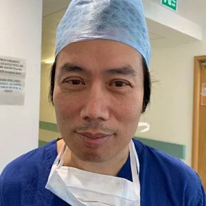 A man wearing medical scrubs, a blue surgical cap, and a face mask lowered around his neck, standing in a hospital corridor.