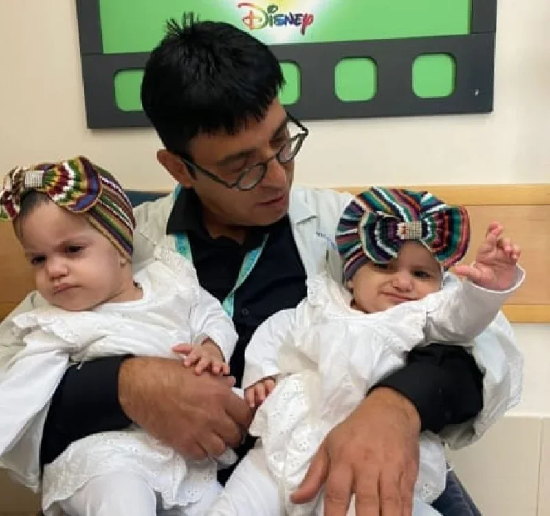 A man holding two young children, both wearing colorful headbands, in a room with a green Disney sign in the background.
