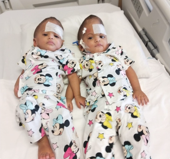 Two young children lying in a hospital bed, wearing matching Minnie Mouse pajamas, with bandages on their heads and hospital monitors nearby.