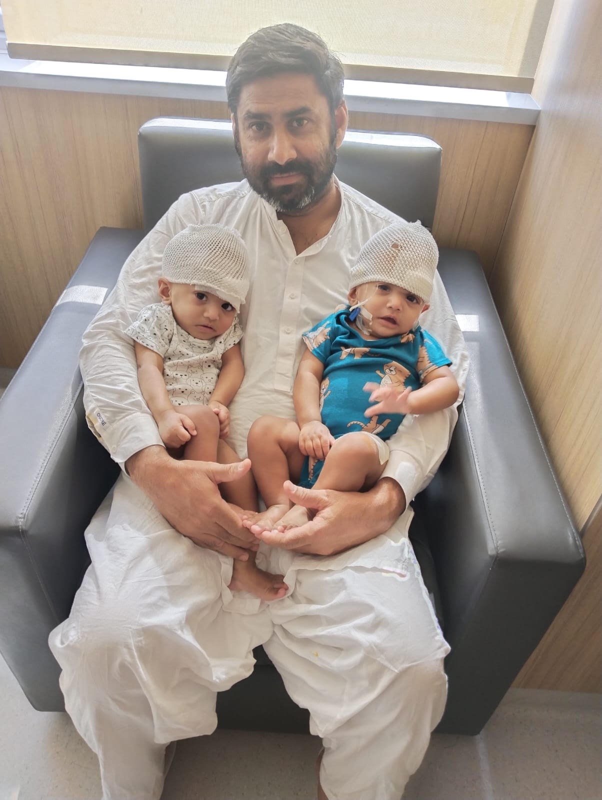 A man with dark hair and a beard sitting on a hospital bed, holding two young children, each wearing a white head cap, with the children sitting on his lap. The children have one child in a patterned shirt and the other in a blue outfit with animal prints.