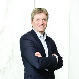 A smiling man in a dark suit with crossed arms, standing against a white background.