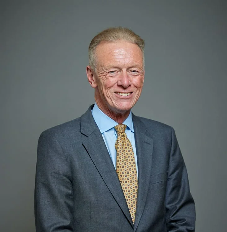 Older man in a dark suit, light blue shirt, yellow patterned tie, smiling, gray background.