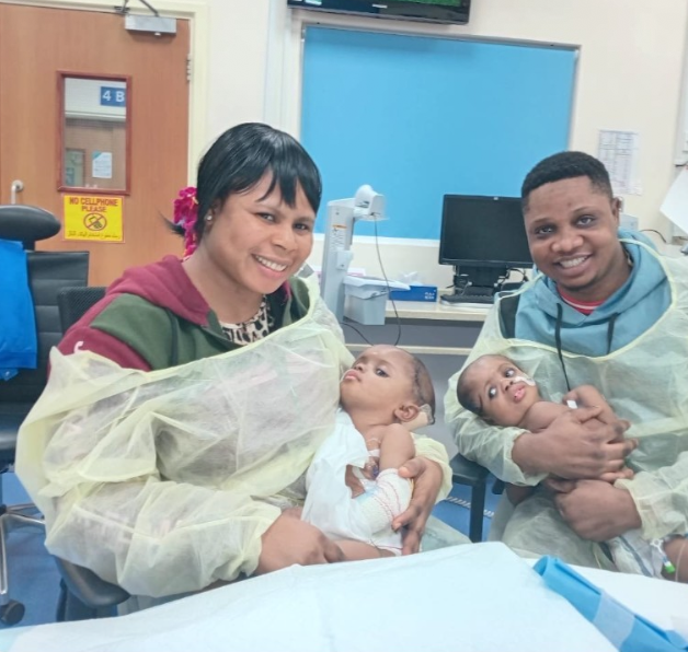 A woman and a man, both wearing yellow hospital gowns, sitting in a hospital room holding two babies. The woman is smiling and holding one baby, and the man is smiling and holding the other baby. There are medical equipment and a computer in the background.