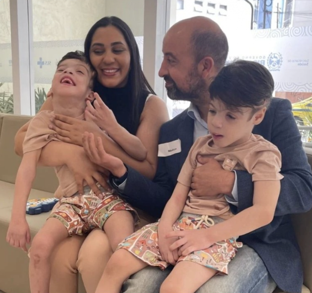 Family of four, including two children with medical conditions, sitting together in a waiting room, smiling and holding each other.