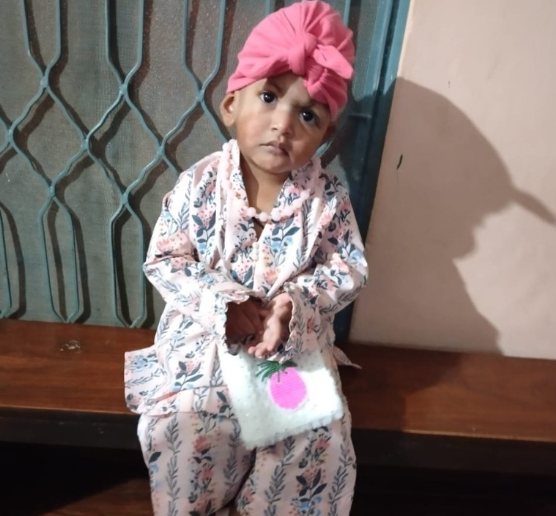 A young girl wearing a pink headwrap and floral pajamas, standing indoors, holding a small bag with a pink circle on it, with a surprised or curious expression.