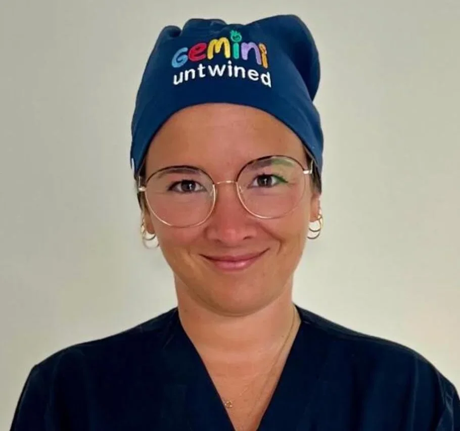 A woman wearing glasses and blue medical scrubs, smiling, with a blue headcover that has colorful text reading 'Gemini untwined'.