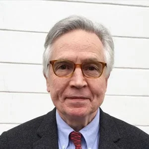 A middle-aged man with gray hair and glasses, wearing a dark suit, light blue shirt, and red tie, standing in front of a white wall.