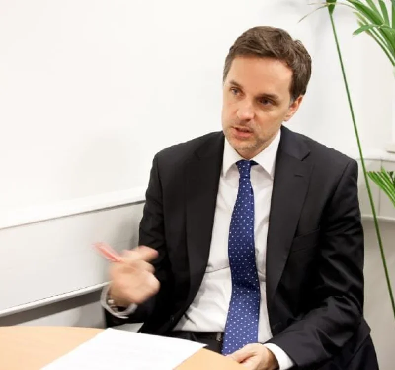 A man in a black suit and blue polka dot tie sitting at a table in an office, talking, with a large green plant in the background.