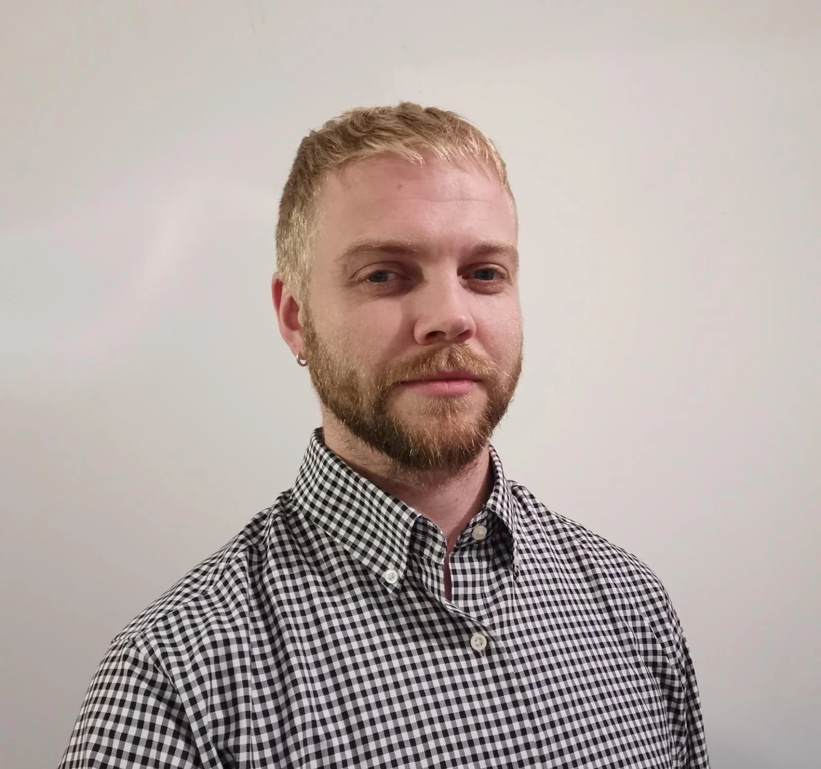 A man with light reddish hair and a beard wearing a black and white checkered shirt, standing against a plain white wall.