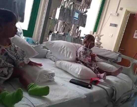 Two children in hospital beds, one with a nasal cannula, in a bright hospital room with medical equipment and a window.
