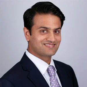 A professional man in a dark suit, white shirt, and tie, smiling against a plain gray background.