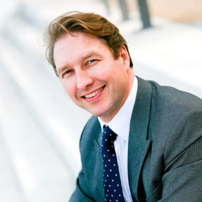 A smiling man with light brown hair, wearing a dark suit, white shirt, and navy blue tie with white polka dots, outdoors on a bright day.